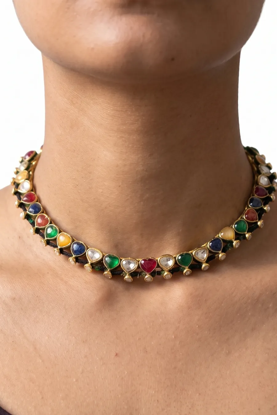 A gold navratna hasli worn at the collarbone — heart-cut ruby, emerald, sapphire and topaz with kundan between them, strung on a dark cord.