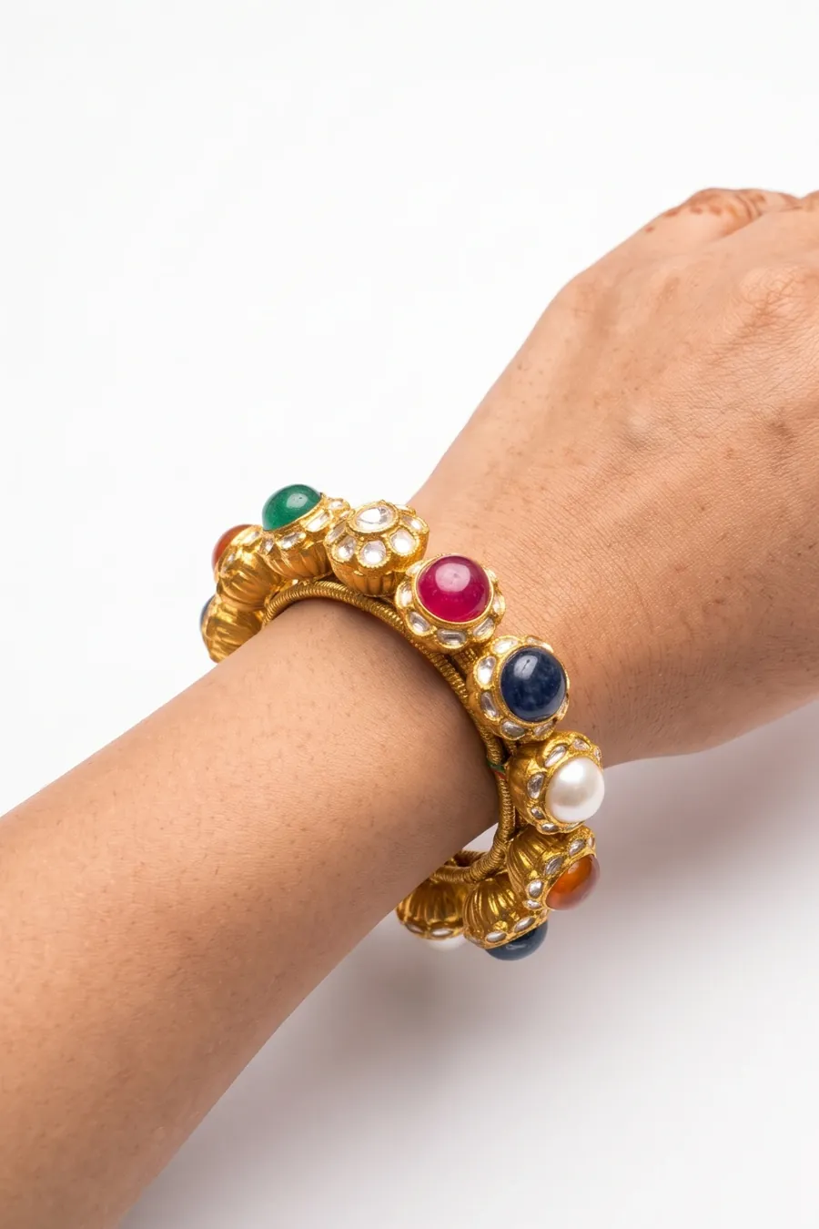 A gold navratna ponchi worn on the wrist — ruby, sapphire, emerald and pearl cabochons in kundan settings between fluted gold caps.